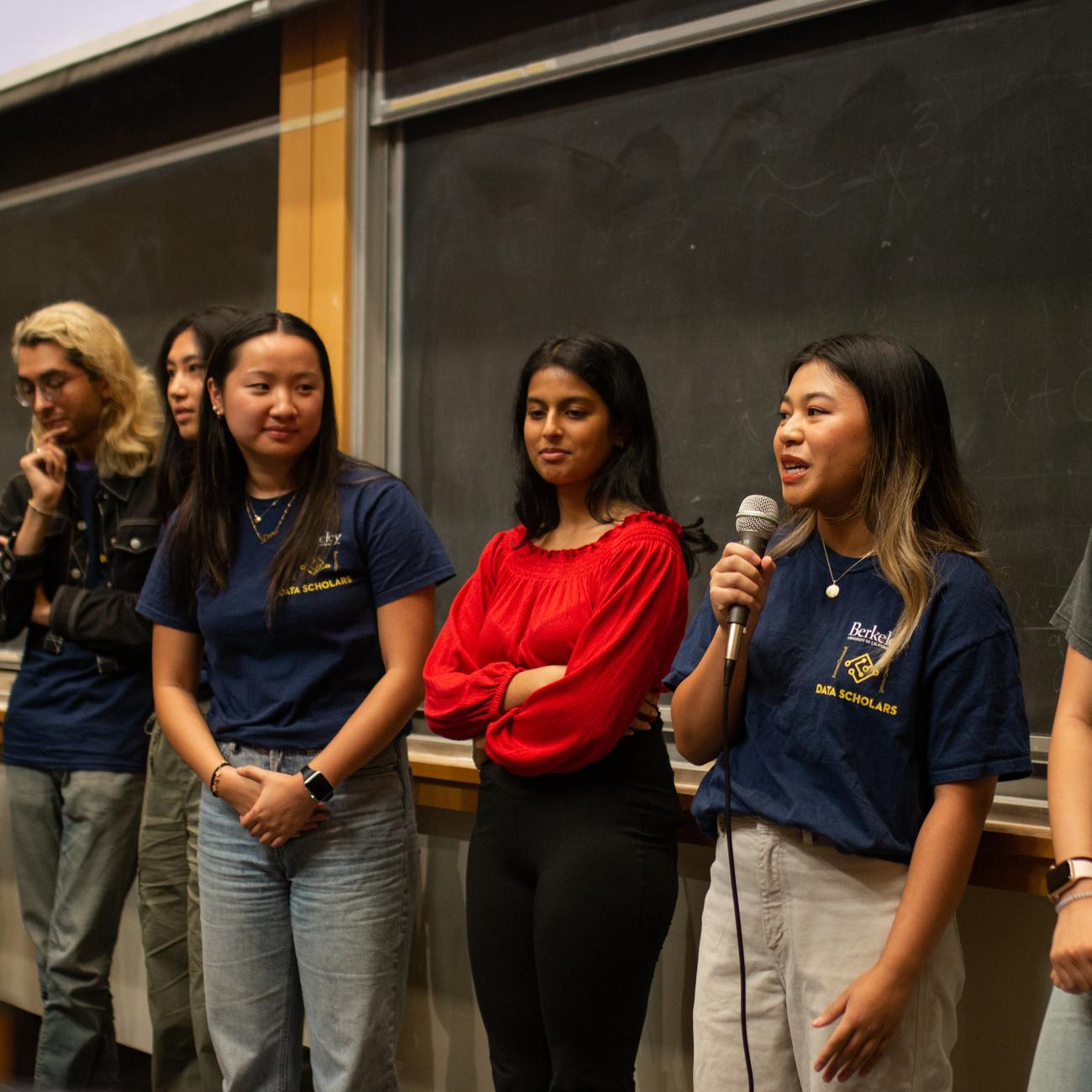 UC Berkeley students standing in a row at the front of a classroom, one student is speaking into a wired microphone