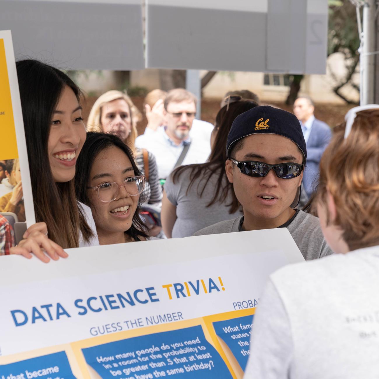 Smiling students at CalDay showing a Data Science Trivia poster to an interested passerby