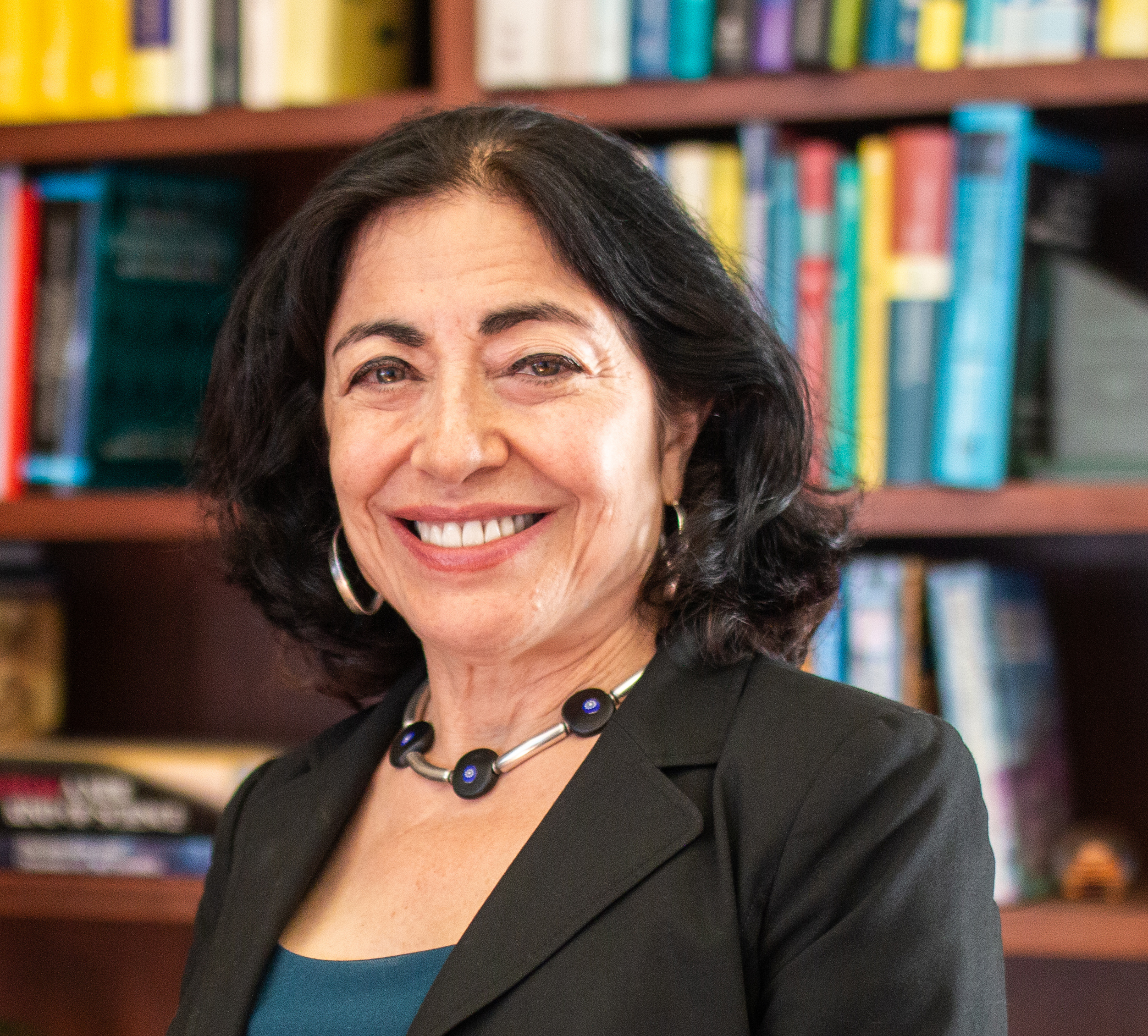 Portrait of Dean Jennifer Chayes wearing a black suit and teal dress, facing the camera and standing in front of a bookshelf